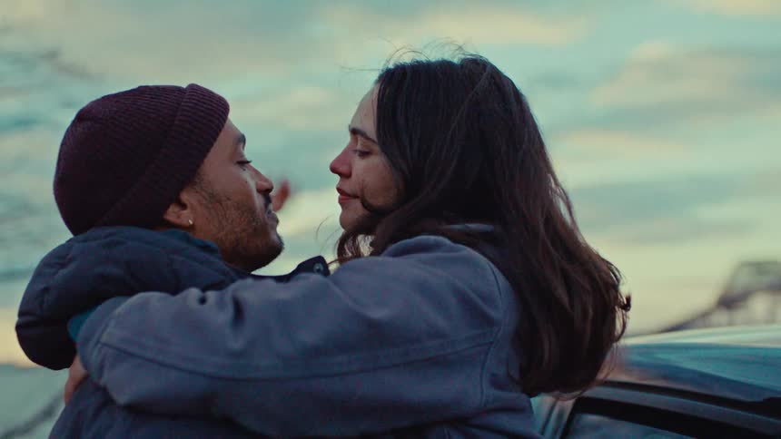 A couple embraces affectionately beside a parked car gazing into each others eyes with a bridge in the background