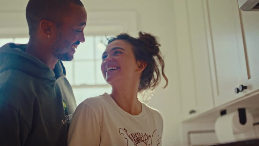 A man and a woman in a brightly lit kitchen smile and kiss intimately with sunlight streaming through a window behind them
