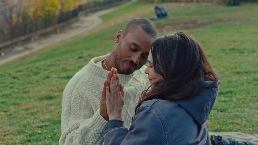 A young couple sits on a grassland in a public park at midday holding hands and laughing together while wearing jackets