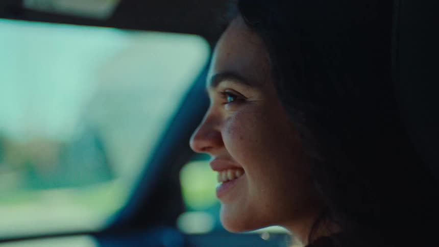 A young woman sits in the passenger seat of a car at midday smiling at the driver as the car moves along the road