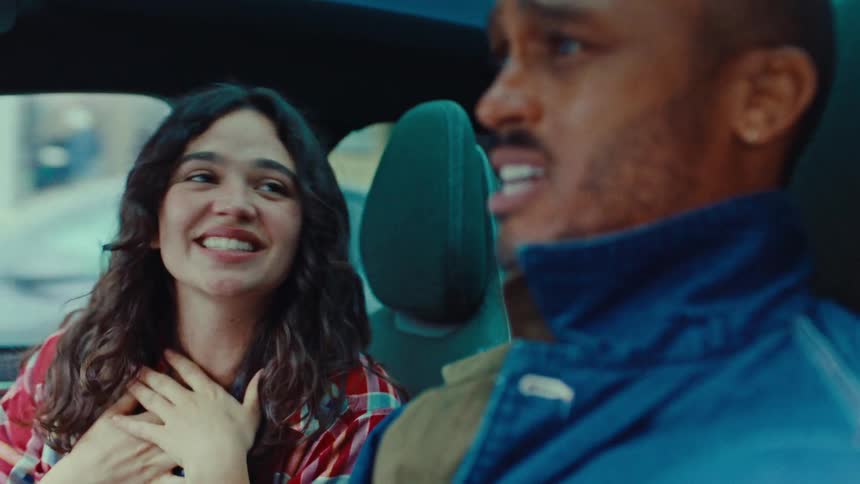 Two people a woman in the passenger seat and a man driving are filmed from a slightly low angle inside a car the woman is animated and smiling as she speaks while the man appears to be listening or joining the conversation