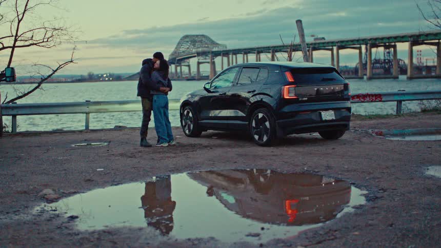 Couple hugging by the water next to a parked car with a bridge in the background