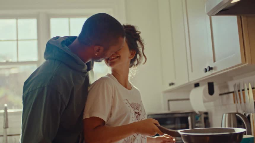In a sunlit kitchen a couple is standing close together by the counter the man wearing a hoodie is embracing the woman who is holding a frying pan and they share a brief kiss while smiling at each other