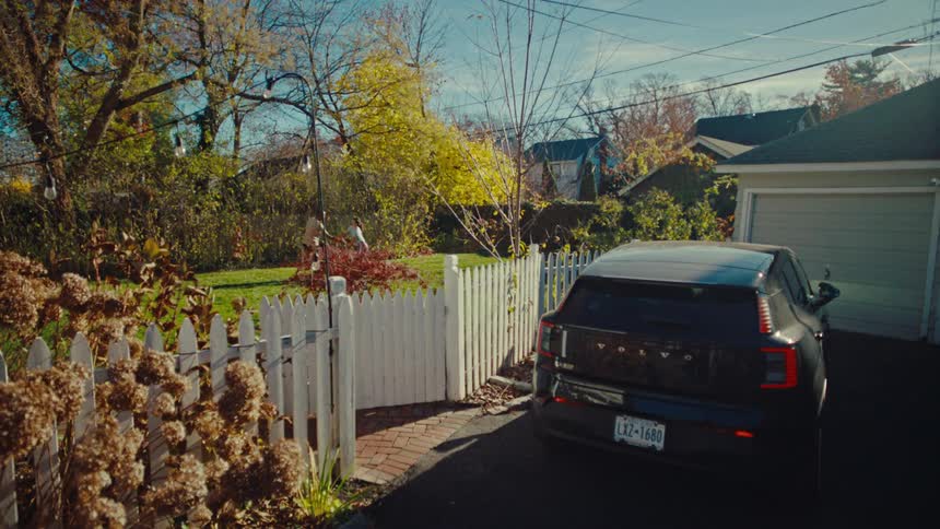 A dark suv is parked in a suburban driveway next to a white picket fence while two people play with a ball in the sunny backyard with a lot of trees surrounding the area
