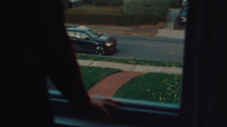 A person in a dark shirt looks out a window onto a suburban street where a black car is parking on the street in front of a green lawn and sidewalk