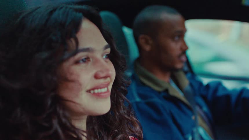 A young couple a black man and a white woman sit inside a car as he drives around the street at midday