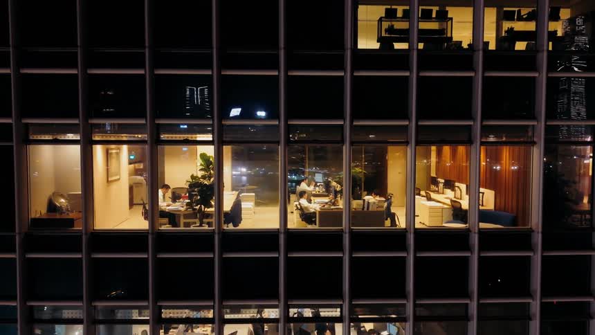 Through a grid of windows the glow of a bustling office at night reveals people working late their screens and desks illuminated against the dark
