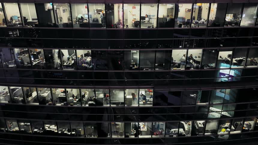 An exterior wide shot captures a modern multistory office building at night the many illuminated windows reveal numerous workers seated at their desks toiling away late in the evening