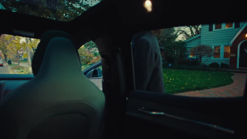 A view from the back seat of a modern car shows a man and a woman walking toward a suburban brick house before the man turns to open the car door