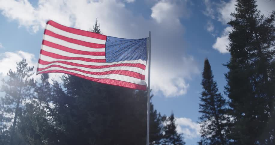 United states flag waving on a pole in front of trees and a partly cloudy sky