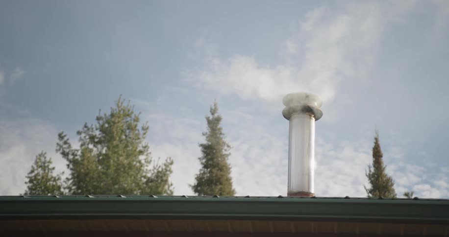 Metal chimney releasing white smoke under blue sky with trees in the background suggesting cozy setting