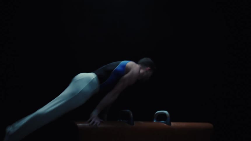 A young athletic man performs gymnastics on wooden flooring inside a dark room