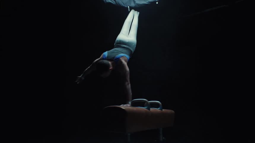 Gymnast in blue and white uniform performs handstand on pommel horse under dramatic spotlight