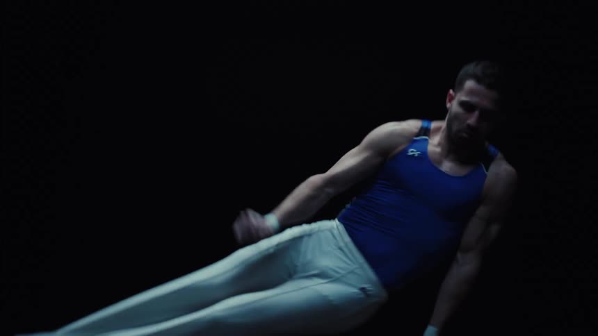 Male gymnast performs a pommel horse routine in a dark area lit by a spotlight