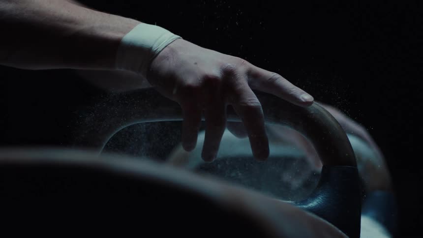 This video is a closeup dimly lit shot of a gymnasts hand chalking up the handles of a pommel horsea hand wrapped with white athletic tape around the wrist firmly places chalk onto the wooden handle of the apparatus causing a cloud of white powd