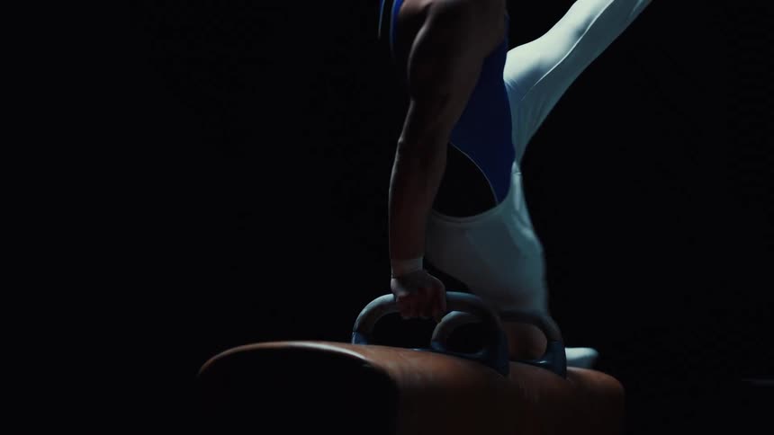 This video features a gymnast performing a routine on the pommel horse in a dimly lit settingthe camera captures the precise controlled movements focusing on the athletes extended legs and powerful upper body as they transition between positions on