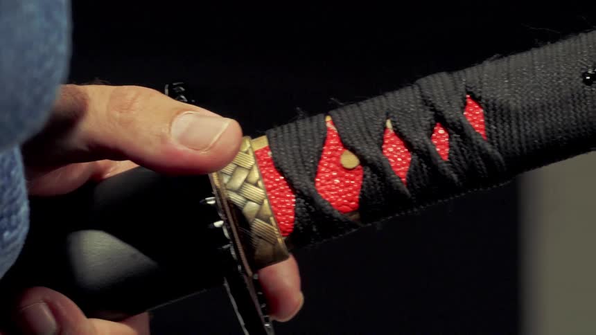 Closeup of a hand gripping the black cord wrapped katana hilt with decorative fittings