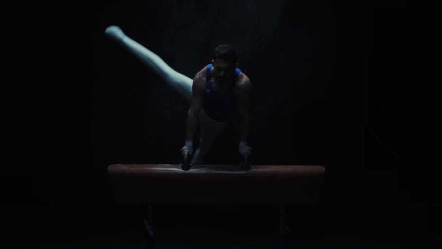 This video shows a male gymnast performing a routine on a pommel horse in a dark dramatically lit setting he executes various complex leg movements and rotations with precision showcasing his strength and skill