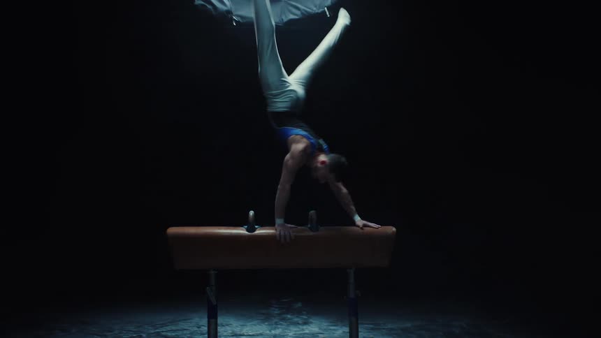 A male gymnast performs a handstand on a pommel horse in a dark gymnasium