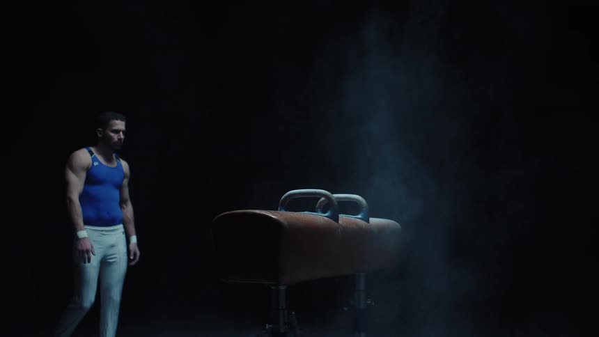 Male gymnast approaches a pommel horse in an area lit by a spotlight