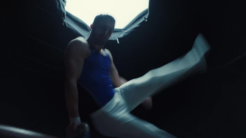 The video shows a gymnast performing on a pommel horse in a dark setting illuminated by a strong overhead light sourcethe athlete is wearing a blue top and white pants executing a dynamic leg movement over the apparatus