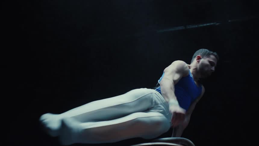 A male gymnast in a blue and white uniform performs a difficult routine on the pommel horse rapidly swinging his legs with precision under a dramatic spotlight in a dark room