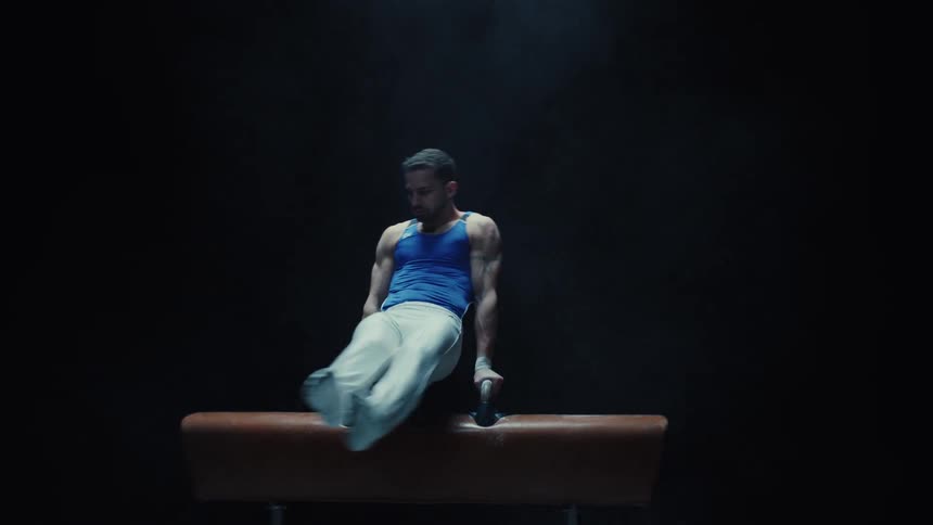 A male gymnast wearing a royal blue tank top and white trousers performs a routine on a pommel horse in a dark spotlightlit settinghe executes a series of coordinated leg movements and circular motions maintaining an elevated position and strong