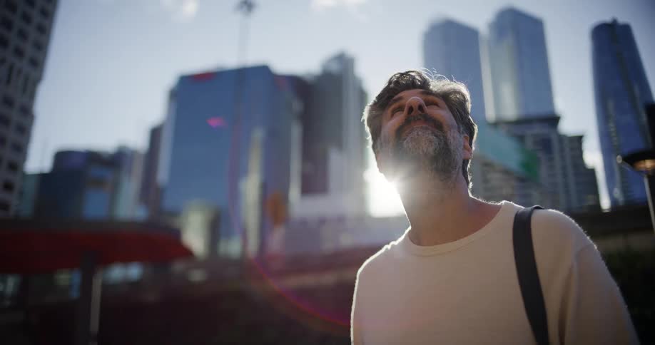 Grayhaired bearded man gazes at modern skyscrapers in urban setting under soft sunlight