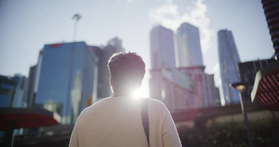 This video shows a person walking away from the camera in a city setting on a sunny daythe person is looking up at several tall modern glass skyscrapers the sun is directly behind the person creating a bright lens flare and backlighting their