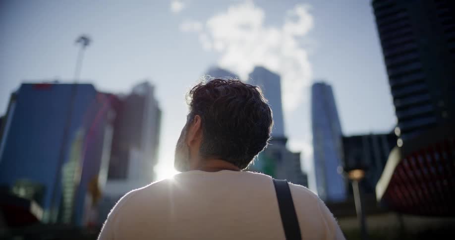Person in white shirt gazes at modern skyscrapers under partly cloudy sky with sun flare
