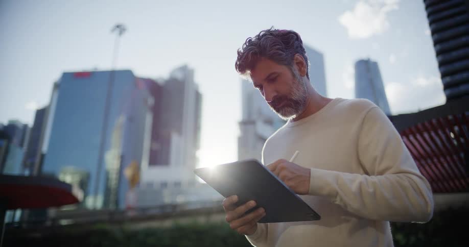 Man stands outside downtown working on a tablet computer with towers and skyscrapers in the distance