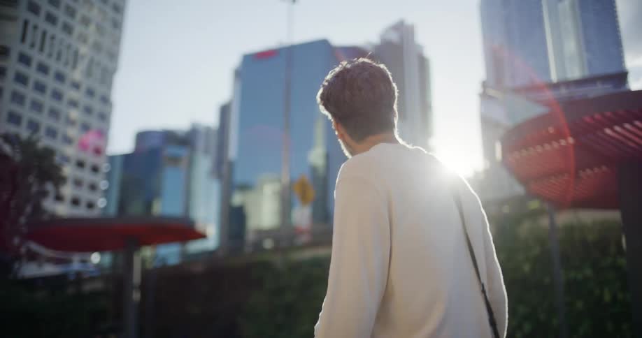 A man with dark curly hair wearing a lightcolored possibly offwhite crewneck sweater and a shoulder strap is filmed from behind and slightly to the sidehe is walking through a sunlit urban area looking toward a cluster of modern glass skyscrap