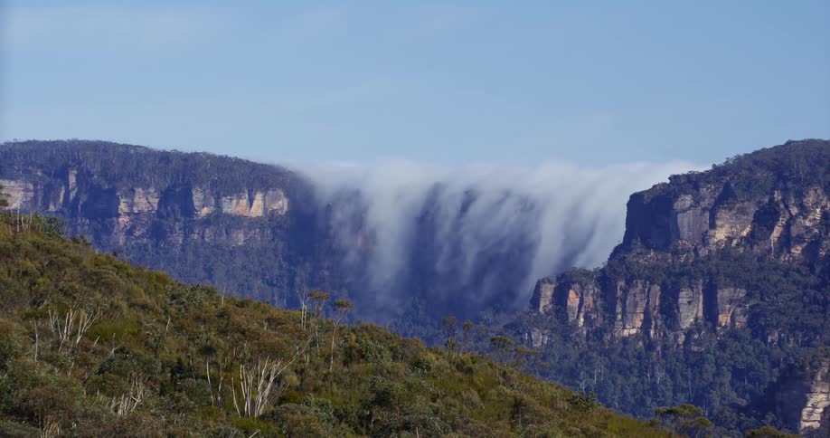 A wide shot of a deep mountain gorge on a sunny day shows steep forested cliffs on either side with clouds or mist flowing and rising from the valley floor between the towering rock formations