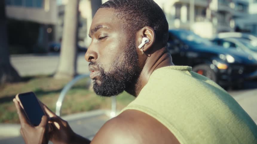 A man sits outside on a sunny street listening to audio through wireless earbuds with a focused expression as he holds his smartphone
