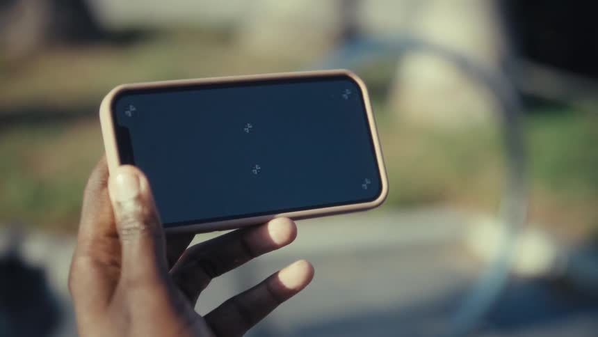 A closeup shot shows a hand holding a smartphone horizontally with a lightcolored case outdoors in daylight while a man walks with a dog in the blurry background