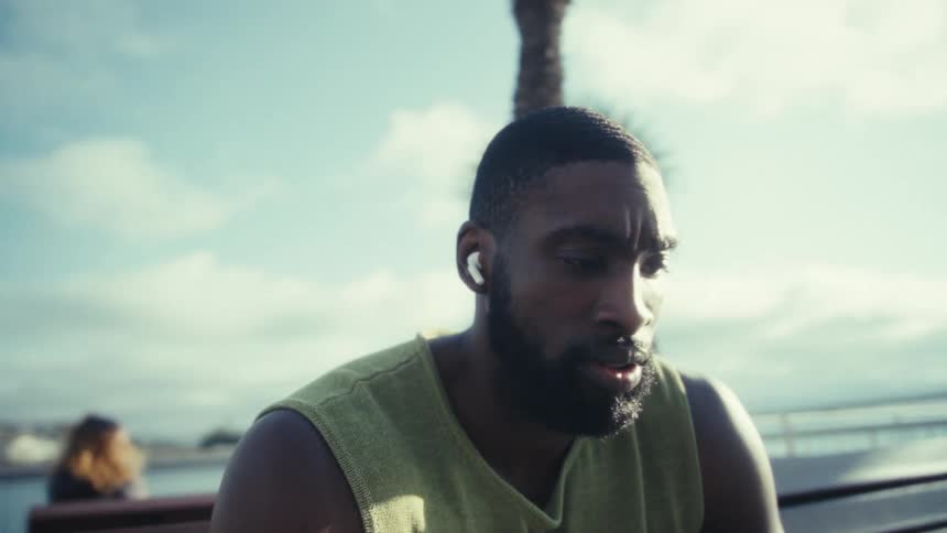 A fit bearded man wearing a yellowgreen tank top and white wireless earbuds is seated outdoors on a wooden bench intently focused on the screen of a lightbrown smartphone held in his hands with a clear blue sky and a water way in the background
