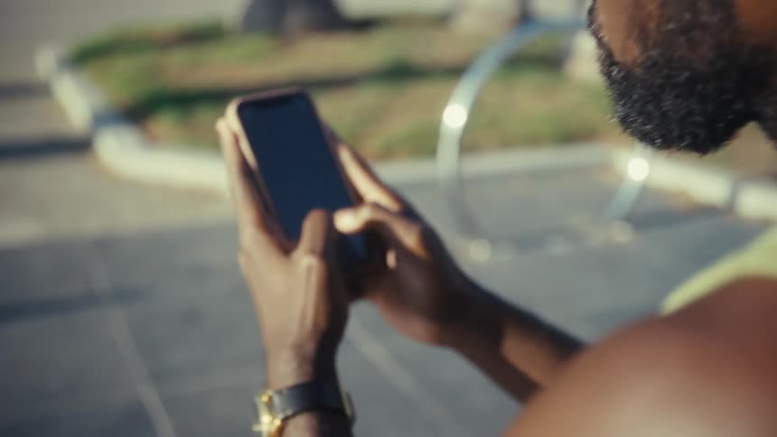 A closeup overtheshoulder shot focuses on a man wearing wireless earbuds and a green tank top who is intensely scrolling and tapping on his smartphone while sitting outdoors on a sunny day