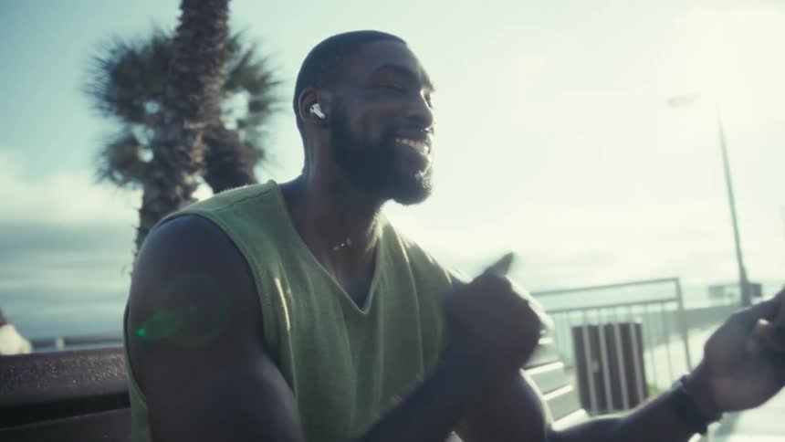 A young black male sits on a public park bench at midday wearing earbuds and holding his smartphone looking excited at the content he is watching