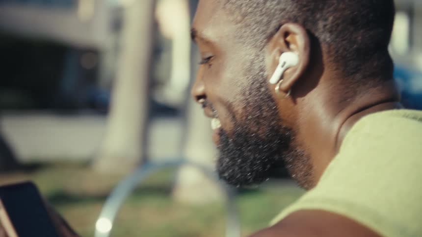 A black man smiles while listening to earbuds and typing on his smartphone outdoors