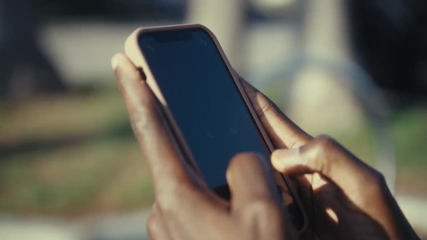 Young black man sitting on a park bench texting on a mobile phone with an urban street on the background outdoors during day time