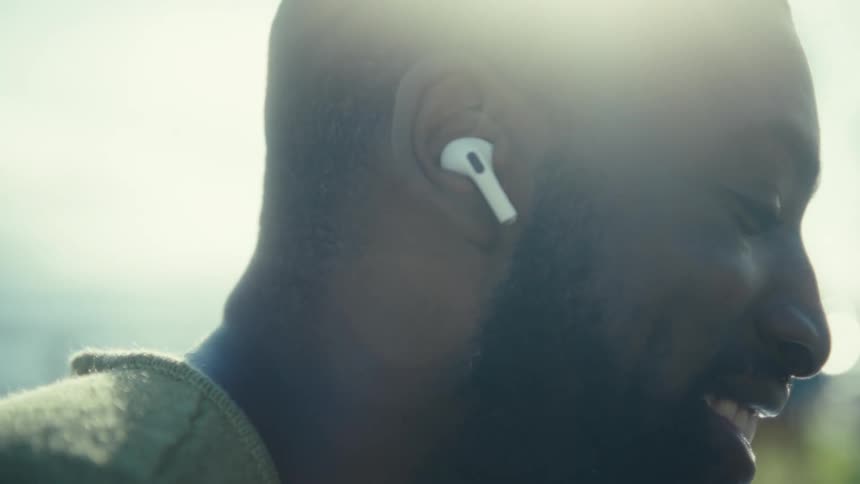 Man smiles while sitting outdoors near the coast using earbuds on a sunny day