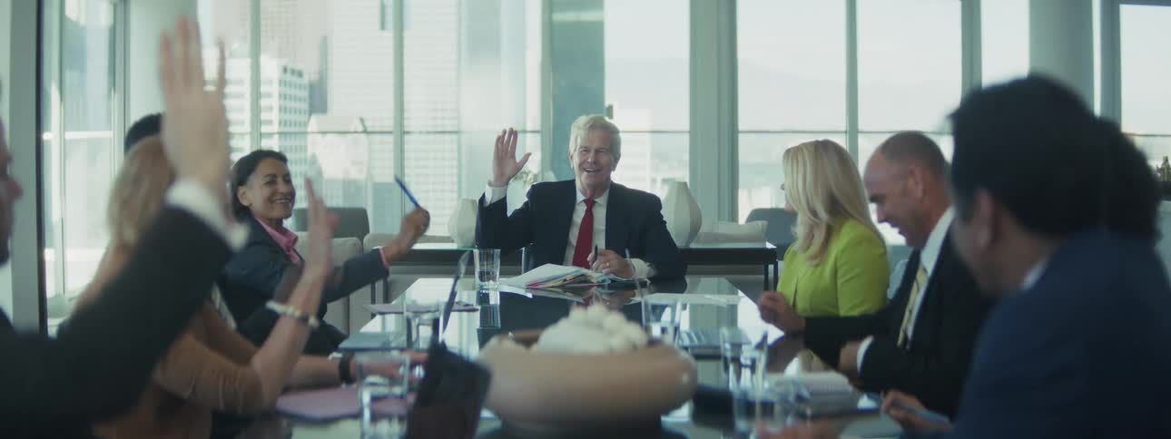 A man in a suit is sitting at a large conference table smiling and waving his hand other people around the table are also raising their hands or laughing during what appears to be a formal meeting