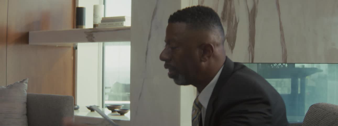 A closeup shot shows a black man in a business suit speaking passionately and gesturing with a document in his hand he looks engaged in a formal conversation within a bright modern office setting