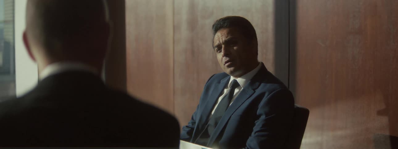 A dramatically lit overtheshoulder shot features a man in a dark suit sitting in a woodpaneled room maintaining a serious focused expression during a formal conversation with an unseen person