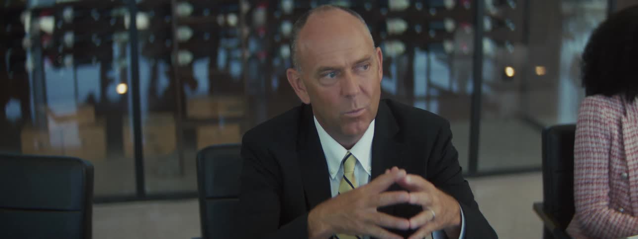 A distinguished older man in a black suit white shirt and a yellowandnavy patterned tie is seated at a conference table with his hands clasped addressing people outside the frame in a modern office with glass walls and reflective lighting fixtures