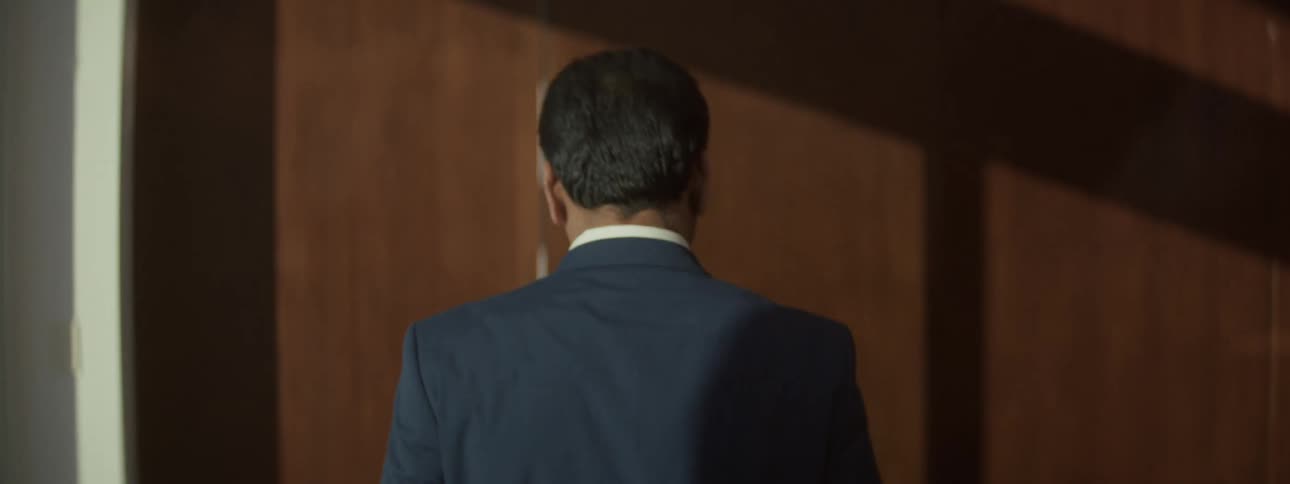 Filmed from behind a man in a blue suit walks through a dimly lit woodpaneled space then turns to the side with a professional smile and opens the door