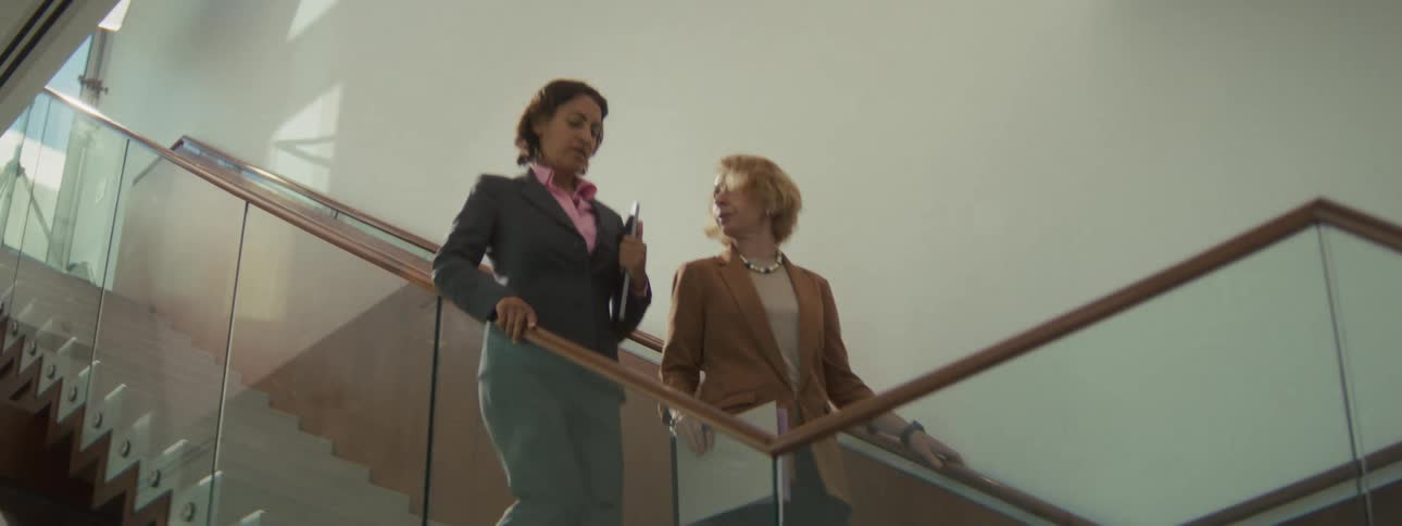 A lowangle shot shows two professional women in business attire descending a modern glass and wood staircase engaged in a focused conversation while carrying documents in a brightly lit building