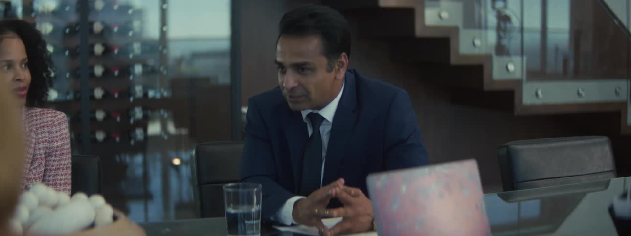 A businessman in a dark blue suit and white shirt sits at a large conference table in a modern office speaking with colleagues while using hand gestures to emphasize his points during a serious discussion