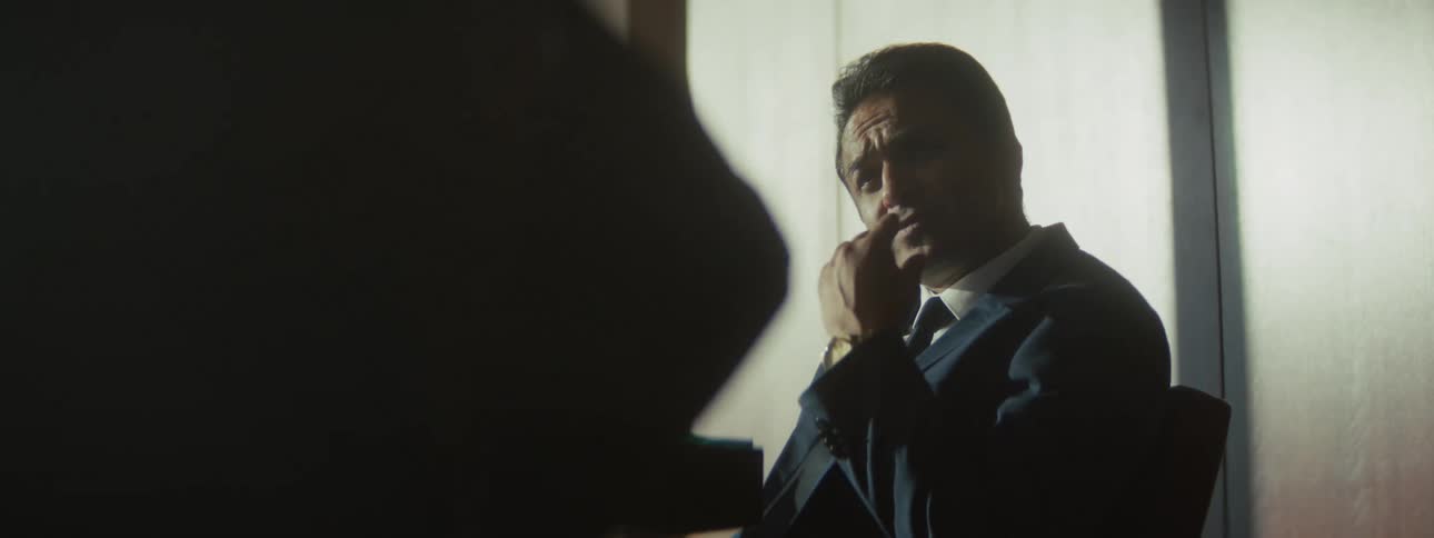 Adult man wearing a suit and tie sitting down and talking to another person in a business meeting indoors during day time