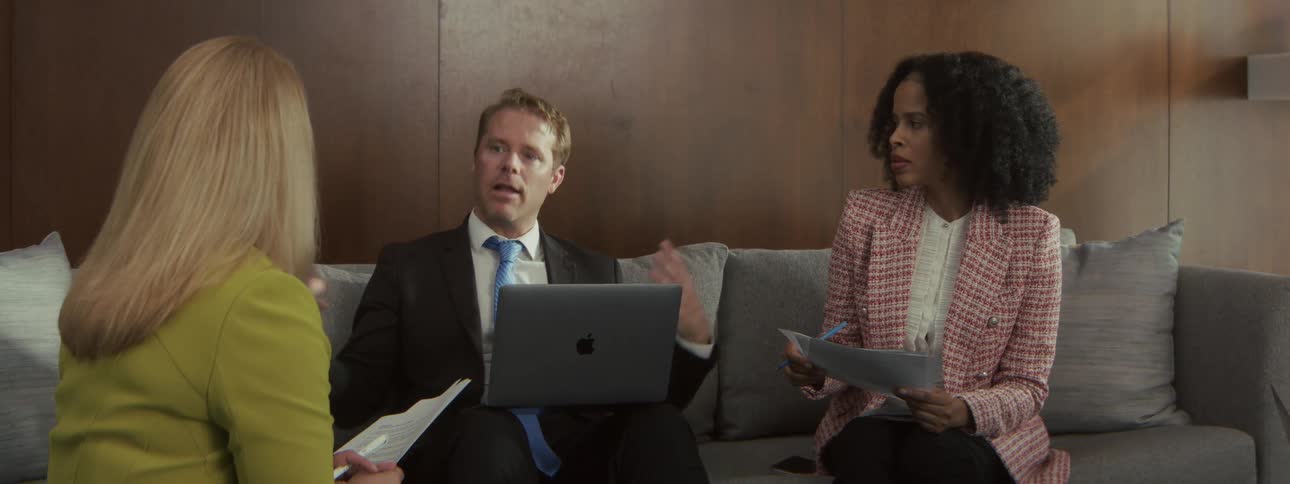 Young adult man sitting down on a couch with a laptop computer on his lap talking to two woman of different ethnicities in a business meeting indoors during day time